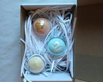 May include: A gift box containing three bath bombs in shades of peach, blue, and yellow. The bath bombs are nestled in white paper strips. The box is open, revealing the contents, and is made of cardboard.