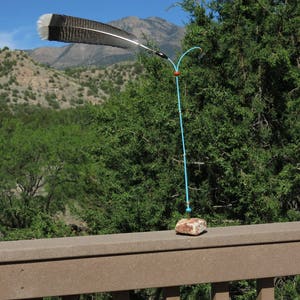May include: A blue metal sculpture with a brown rock base and a gray and white feather attached to the top. The sculpture is outdoors with a mountain range in the background.