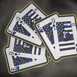 May include: Five playing cards with a black and white American flag design and a blue stripe. The cards are all King of Spades and have the text "BET WE'LL FIND YOU" on them. The cards are arranged in a fan shape.