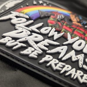 May include: A black rectangular morale patch featuring a raised white graphic design with the text FOLLOW YOUR DREAMS BUT BE PREPARED. The design includes a colorful rainbow arching over a figure wearing a red and black plaid shirt.