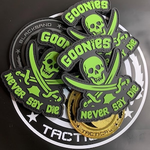 May include: A collection of black and lime green rubberised morale patches featuring a skull and crossbones design with the text GOONIES and NEVER SAY DIE. The patches are layered over a circular black and white tactical logo and a gold coin.