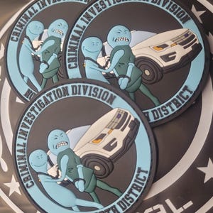 May include: Three round black and blue patches with white lettering that reads "Criminal Investigation Division" and "North District". Each patch features a cartoon image of two blue figures in handcuffs being escorted by a police car.