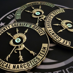 May include: Three gold-colored sheriff's badges with black backgrounds. Each badge features a snake wrapped around a pool ball with the number 8, and three arrows pointing towards the ball. The text on the badges reads "Lee County Sheriff's Office" and "Tactical Narcotics Team".