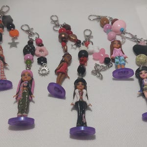 Könnte beinhalten: Eine Sammlung handgefertigter Schlüsselanhänger mit kleinen Puppenfiguren. Jeder Schlüsselanhänger hat ein einzigartiges Design mit bunten Perlen, Charms und einem Metallverschluss. Die Puppen sind in verschiedenen Outfits gekleidet und stehen auf lila Sockeln.