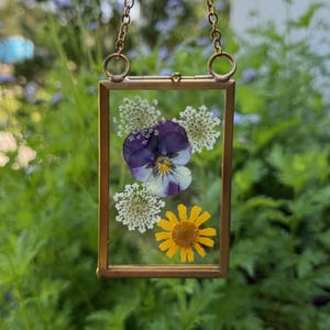 May include: A rectangular, gold-toned framed pendant with pressed flowers. The pendant features a purple and white pansy, a yellow daisy, and white lace flowers. A delicate gold chain suspends the pendant, showcasing a botanical theme.