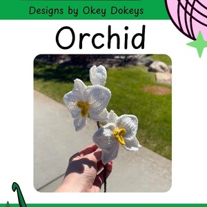 May include: A crochet orchid flower with white petals and a yellow center, attached to a thin brown stem. The image includes the text Okey Dokeys, Designs by Okey Dokeys, and Orchid, along with a pink yarn ball icon and a green crochet hook icon.