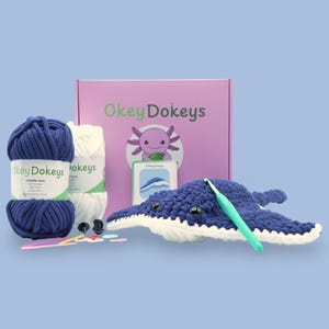 May include: A craft kit featuring a blue and white crocheted manta ray, a pink box with the text "Okey Dokeys", and two skeins of yarn. The kit includes a crochet hook, safety eyes, and other crafting supplies. The overall color scheme is blue, white, and pink.