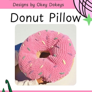 May include: A pink crocheted donut pillow with colorful sprinkles. The pillow has a hole in the center and is labeled "Donut Pillow" with "Designs by Okey Dokeys" above. A crochet hook is in the lower left corner.