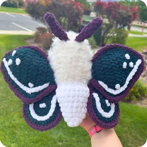 May include: A handcrafted crochet moth plush featuring a fuzzy white body and two large wings. The wings are dark teal with white accents and a deep purple border. Two dark purple antennae extend from the top of the head, creating a soft texture.
