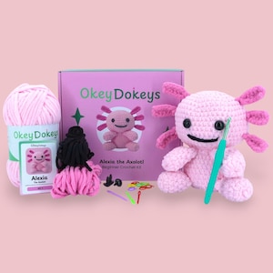 May include: A beginner crochet kit featuring a pink box labeled OkeyDokeys Alexia the Axolotl, a skein of light pink yarn, a finished pink crocheted axolotl plush, a green crochet hook, black safety eyes, colorful stitch markers, and a needle.