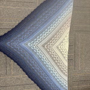 May include: A handmade, triangular shawl in shades of blue, from deep navy to light blue and white. The shawl features an open, lacy crochet design with intricate patterns. The shawl is made of soft yarn and is perfect for cool weather.