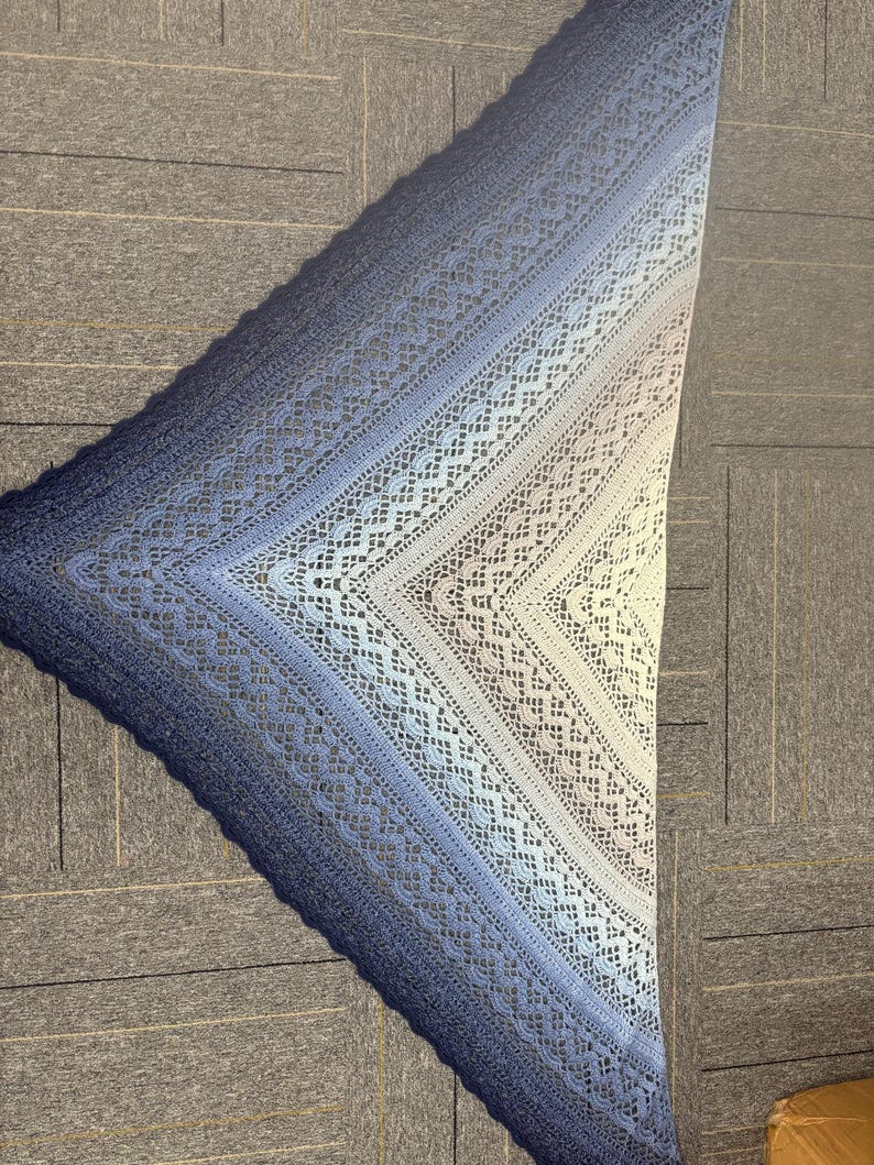 May include: A handmade, triangular shawl in shades of blue, from deep navy to light blue and white. The shawl features an open, lacy crochet design with intricate patterns. The shawl is made of soft yarn and is perfect for cool weather.