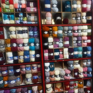 May include: A wall of shelves filled with colorful yarn balls. The yarn is organized by color and brand. The yarn is in various shades of red, blue, green, yellow, and purple. The yarn is labeled with the brand name "NAKO".