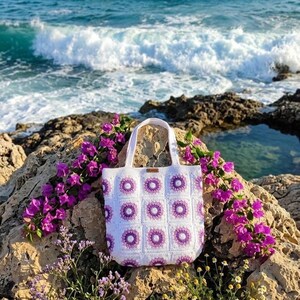 May include: A white crochet tote bag with a purple floral pattern. The bag has a rectangular shape with a handle and is displayed on a rocky surface with purple flowers. The background features the ocean and a bright blue sky.