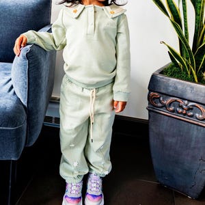 May include: A child's light green outfit, including a long-sleeved top with a collared neckline and matching pants. The outfit is adorned with small floral accents. The child is wearing colorful sneakers with white soles.