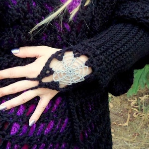 May include: Black crochet fingerless gloves with a silver floral design on the wrist. The gloves are made of a thick, textured yarn. The background is a dark, textured fabric.