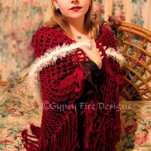 May include: A woman wearing a red crocheted shawl with white fringe. The shawl has a lacy pattern and is draped over her shoulders. The woman is sitting on a floral patterned couch.