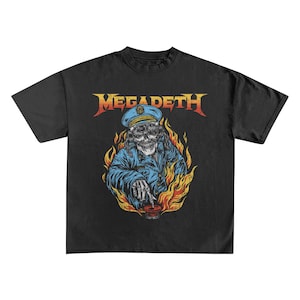 May include: Black t-shirt featuring the band name "Megadeth" in orange and yellow lettering. The graphic depicts a skull in a blue uniform and hat, surrounded by flames. The skull is holding a red object.