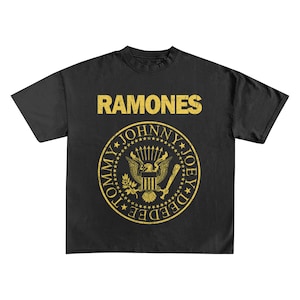 May include: Black t-shirt featuring the band name "RAMONES" in gold block letters above a gold emblem. The emblem includes an eagle, stars, and the names "TOMMY", "JOHNNY", and "JOEY DEE DEE" in a circular design.