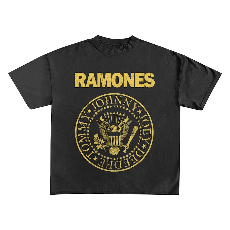 May include: Black t-shirt featuring the band name "RAMONES" in gold block letters above a gold emblem. The emblem includes an eagle, stars, and the names "TOMMY", "JOHNNY", and "JOEY DEE DEE" in a circular design.
