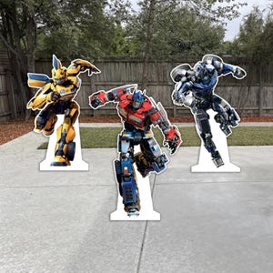 May include: Three cardboard cutouts of Transformers characters: Bumblebee in yellow and black, Optimus Prime in red, blue, and silver, and a third character in blue and silver. The figures are posed in action stances and stand on white bases.