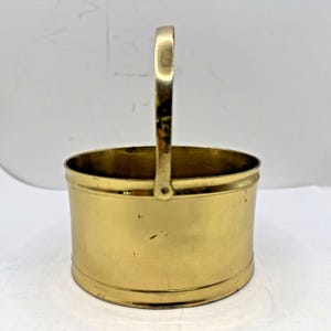 May include: A polished brass bucket with a smooth, cylindrical body and a single upright handle attached with rivets. The metal surface has a reflective gold-toned finish with subtle variations in texture and light across the exterior.