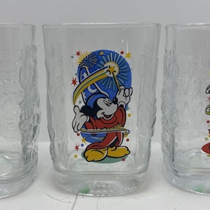 May include: Three clear glass tumblers featuring colorful illustrations of Mickey Mouse. The first shows Mickey pointing upward with fireworks, the second depicts Mickey as a sorcerer with a wand, and the third shows Mickey in safari gear.