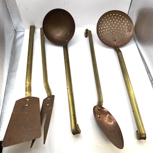 May include: A set of five kitchen utensils featuring copper heads and long brass handles. The collection includes a solid turner, a slotted turner, a deep ladle, a solid spoon, and a skimmer, all with riveted construction and curved handle ends.