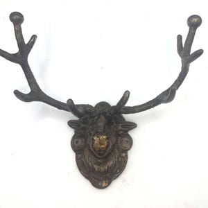 Puede incluir: Un gancho de pared en tono bronce oscuro con forma de cabeza de ciervo y astas prominentes. La cabeza presenta un acabado texturizado con una nariz en tono dorado metálico más claro. Las astas se extienden hacia afuera para colgar objetos.