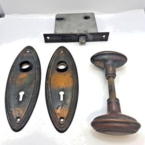 May include: A set of antique door hardware including two oval-shaped brass backplates with keyholes, a matching brass door knob with a square spindle, and a rectangular metal mortise lock mechanism with a latch and a keyhole opening.