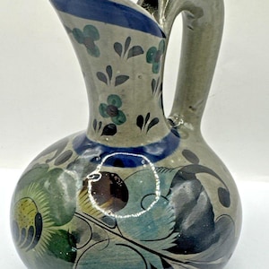 May include: A ceramic jug with a rounded body and a curved handle. The jug is decorated with floral and leaf patterns in blue, green, and black on a grey background. A blue band highlights the rim.