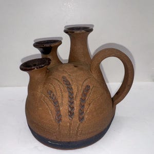May include: A brown ceramic pitcher with three spouts and a handle. The pitcher has a textured surface and is decorated with an embossed wheat design. The base of the pitcher is dark blue.
