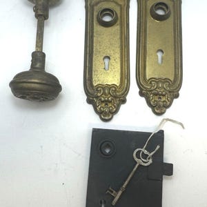 May include: A set of antique door hardware including two brass door knobs connected by a metal spindle, two ornate rectangular brass backplates with keyholes, and a black metal mortise lock box with a single skeleton key attached by a string.