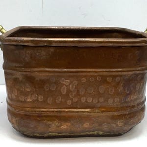 Puede incluir: Un cubo de metal color cobre con dos asas de latón. El cubo tiene forma rectangular con esquinas redondeadas y una superficie texturizada. Las asas son curvas y están unidas al borde superior. Objeto decorativo.