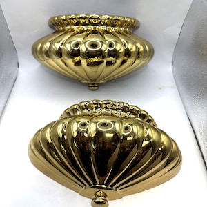 May include: Two matching wall-mounted sconces featuring a polished gold-tone finish. Each fixture has a semi-circular, shell-like design with vertical fluted ridges and a small, rounded decorative knob at the bottom center point.