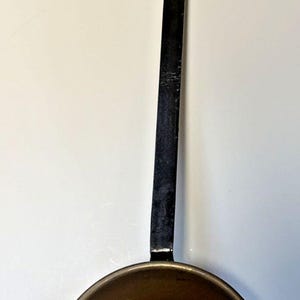 May include: A vintage copper pan with a long, black handle and a small loop at the end. The pan has a rounded bowl shape and a dark, aged patina, suggesting it's been well-used. The handle is approximately 30 cm long.