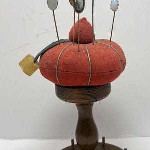 May include: A vintage pin cushion with a red, pumpkin-shaped cushion, adorned with decorative pins. The cushion sits atop a wooden stand with a round base and a turned pedestal. A small, square, yellow object hangs from the side.