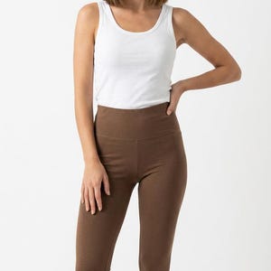 May include: A white sleeveless tank top with a scoop neckline paired with high-waisted solid brown leggings. The leggings feature a wide waistband and a fitted silhouette that extends down to the ankles, creating a simple and clean look.