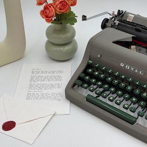 May include: A vintage gray Royal Quiet De Luxe typewriter with green keys sits next to a letter and envelope sealed with red wax. A small vase holds orange flowers. The typewriter has the word "ROYAL" on it.