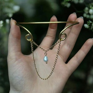 Può includere: Un bracciale dorato con una delicata catena e un singolo pendente di perla. Il bracciale ha un design semplice ed elegante, con una barra curva e volute decorative. La catena pende dal centro, aggiungendo un tocco di raffinatezza.