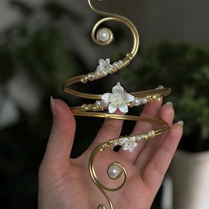 Può includere: Un bracciale dorato con un design a spirale, ornato da accenti floreali bianchi e perle finte. Il bracciale si avvolge attorno al braccio, con dettagli delicati e un'estetica aggraziata ed elegante.