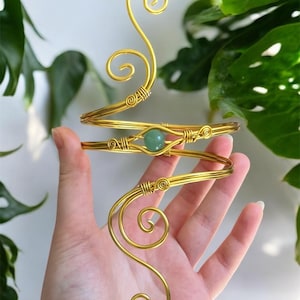 May include: A gold-colored arm cuff with a swirling design and a green gemstone. The cuff wraps around the arm with a spiral design. The gemstone is a small, round, light green bead. The cuff is held in a hand.