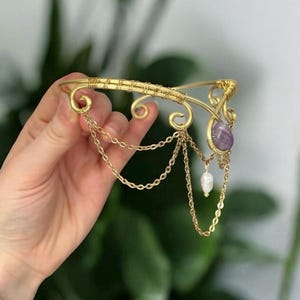 Op de afbeelding: Een goudkleurige, met draad omwikkelde armmanchet met decoratieve krullen, fijne hangende gouden kettinkjes, een enkele ovale paarse amethist edelsteen en een kleine witte traanvormige parel die aan de metalen structuur hangt.