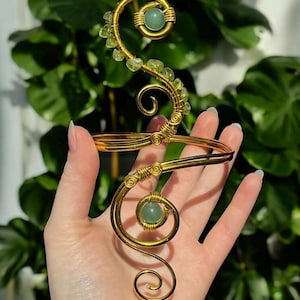 Könnte beinhalten: Ein goldfarbener, drahtgewickelter Armreif mit zwei runden, hellgrünen Steinperlen und einer Reihe kleiner, blassgrüner Kristallsplitter entlang der oberen Spirale. Der Metalldraht ist zu komplizierten, geschwungenen Mustern geformt.