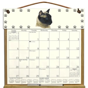 May include: A wooden easel calendar with a cream background and a picture of a Siamese cat. The calendar is for January and features the days of the week and dates. The calendar also includes moon phases and special days, such as Martin Luther King Day.