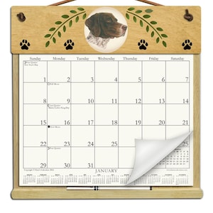 May include: A wooden wall calendar for January featuring a German Shorthaired Pointer illustration at the top. The calendar includes black paw print accents, green leaf motifs, and a white pen resting on the bottom wooden ledge. The calendar grid displays dates, lunar phases, and holidays.
