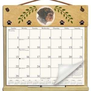 May include: A wooden wall calendar featuring a portrait of a brown and white dog in an oval frame, flanked by green leaf illustrations and black paw prints. The calendar displays the month of January with a curled corner and a silver pen attached.