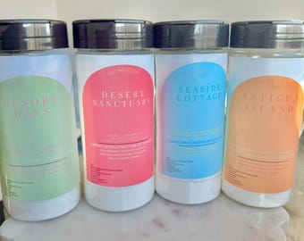May include: Four clear plastic bottles with black lids and colorful labels. The labels have text that reads "Resort Days", "Desert Sanctuary", "Seaside Cottage", and "Antigua Island". The bottles are on a white marble surface with gold bee accents.