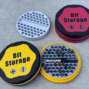 May include: A set of four round bit storage containers. Two containers are closed, with the text "Bit Storage" and a plus and minus symbol on the lids. The open container reveals various drill bits and a DeWalt tool. The containers are black, red, yellow, and gray.