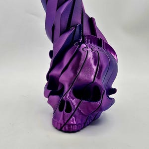May include: A metallic purple sculpture of a skull with a flowing, layered design. The skull's features are detailed, with visible eye sockets and a defined jawline. The sculpture has a glossy finish, reflecting light.